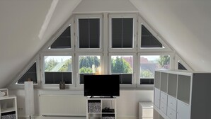 Une pièce avec un plafond en pente, de grandes fenêtres équipées de stores, un téléviseur à écran plat posé sur un meuble, un radiateur et des placards intégrés.