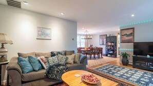 Ein Wohnzimmer mit Sofa, Couchtisch und Fernseher. Es gibt einen Essbereich mit Tisch und Stühlen sowie ein Bücherregal mit Büchern und Dekorationsgegenständen.