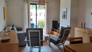 Una sala de estar moderna con un sofá, una mesa de comedor y un televisor. Hay un balcón con vistas a edificios y zonas verdes.