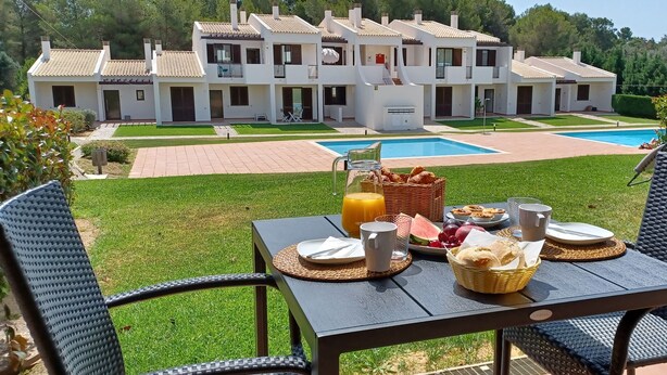 Una mesa de comedor con alimentos para el desayuno, una piscina y un complejo de villas al fondo.