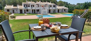 A dining table with breakfast items, a pool, and a villa complex in the background.