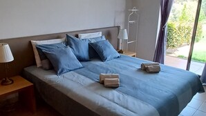 Una cama grande con colcha y almohadas azules, una mesita de noche con lámpara, y una ventana con cortinas.
