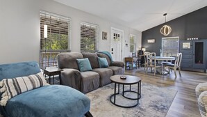 Ein großzügiger Wohnbereich mit grauem Sofa, blauem Sessel und rundem Couchtisch. Es gibt einen Esstisch mit Stühlen, eine Küchenzeile und einen Deckenventilator.