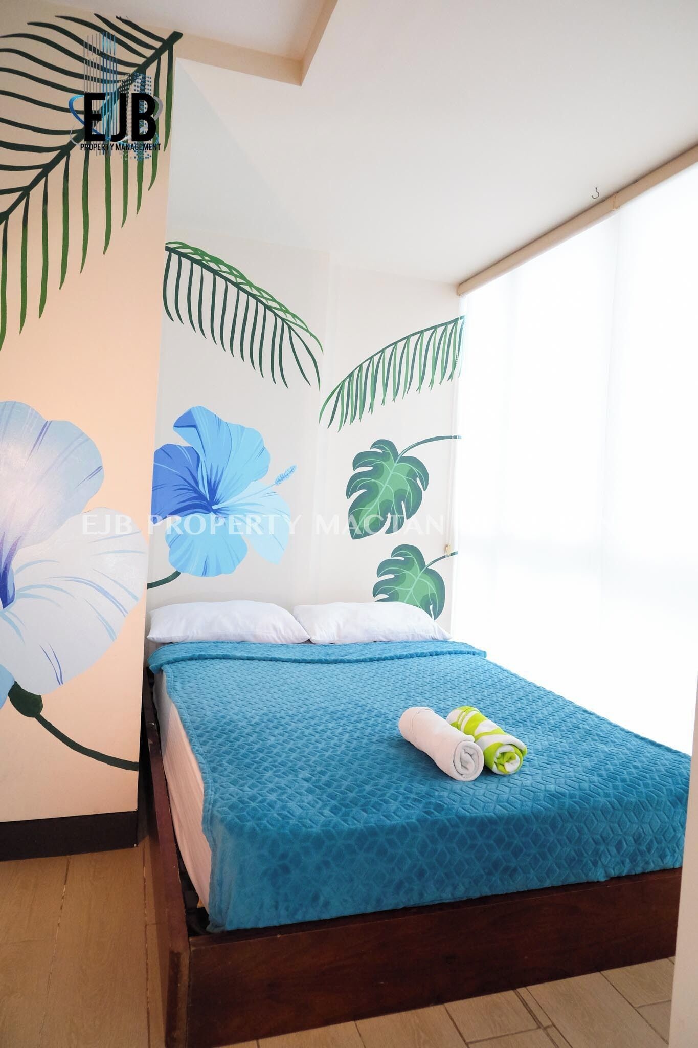 A bedroom with a bed, a blue bedspread, and a wall mural with large flowers and leaves.