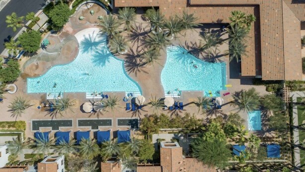 Luchtbeeld van een resort met twee zwembaden, omringd door palmbomen, ligbedden en een kleine waterval.
