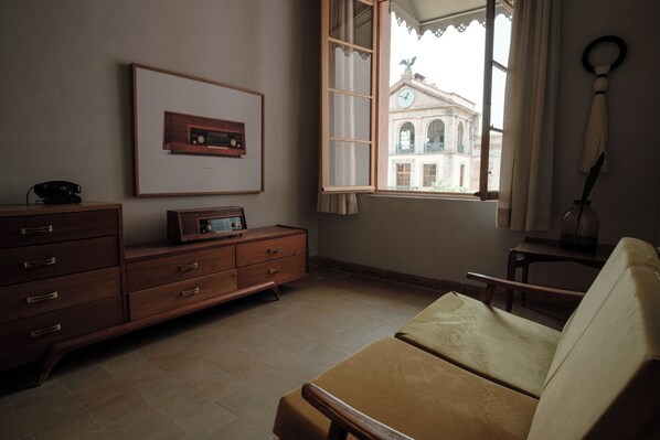 Una habitación con un aparador de madera, un cuadro enmarcado de una radio, una ventana con vista a un edificio y un sofá color beige.