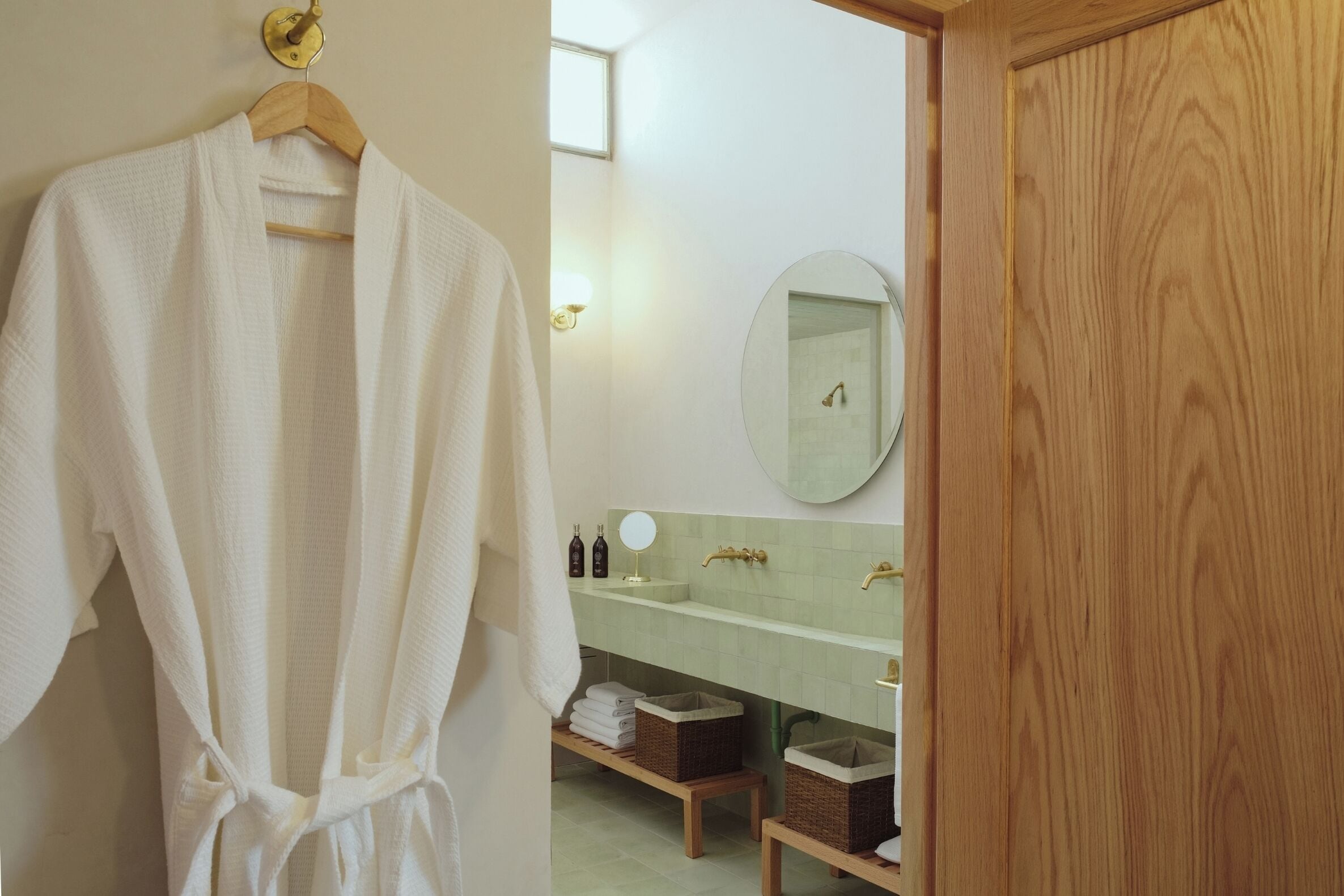 A bathroom with a wooden door, a white robe hanging on a wooden hanger, a green-tiled sink, and a mirror.