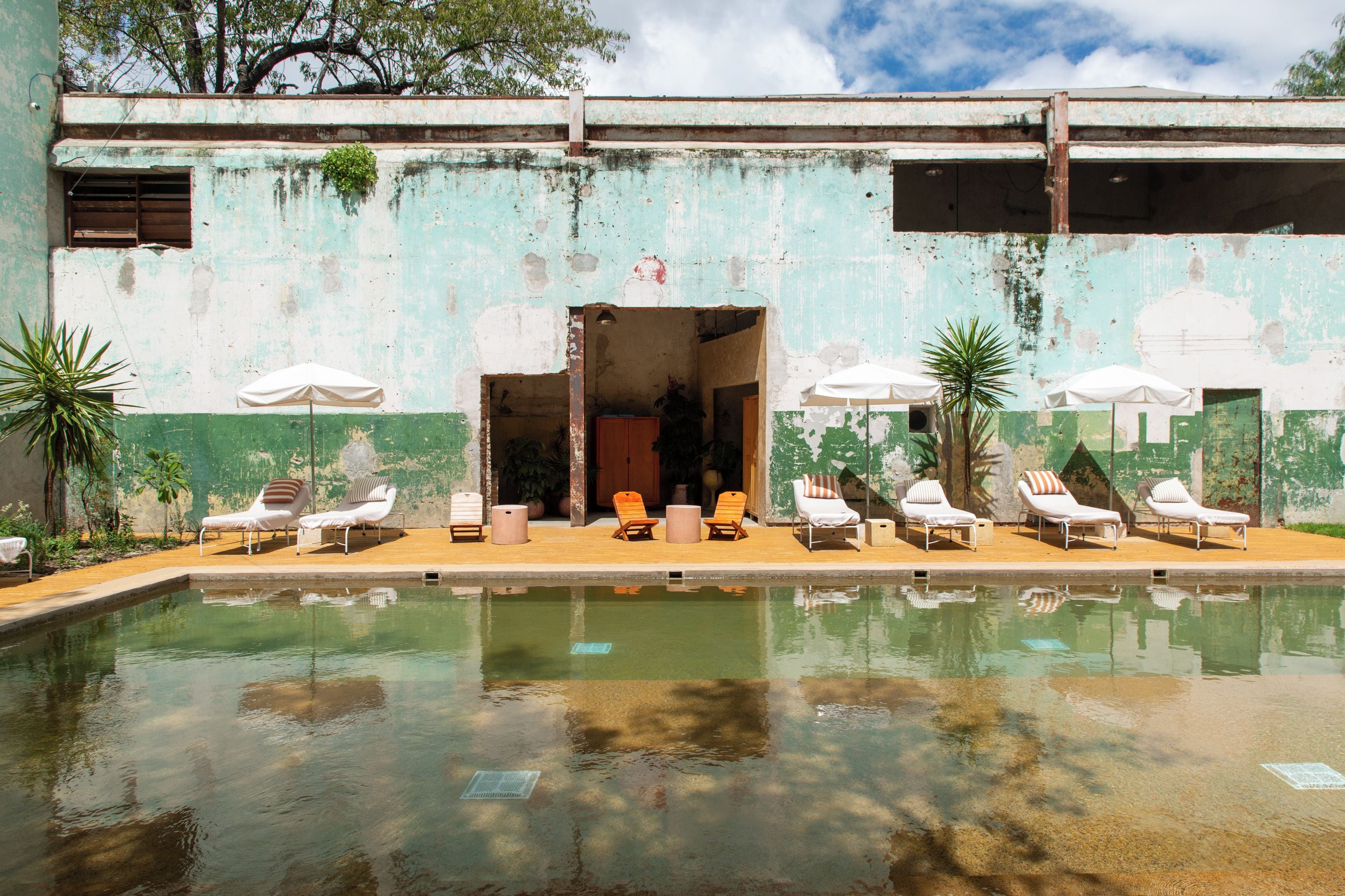 Un área de piscina con sillones de descanso y sombrillas, una fachada de edificio desgastada y un cielo despejado.
