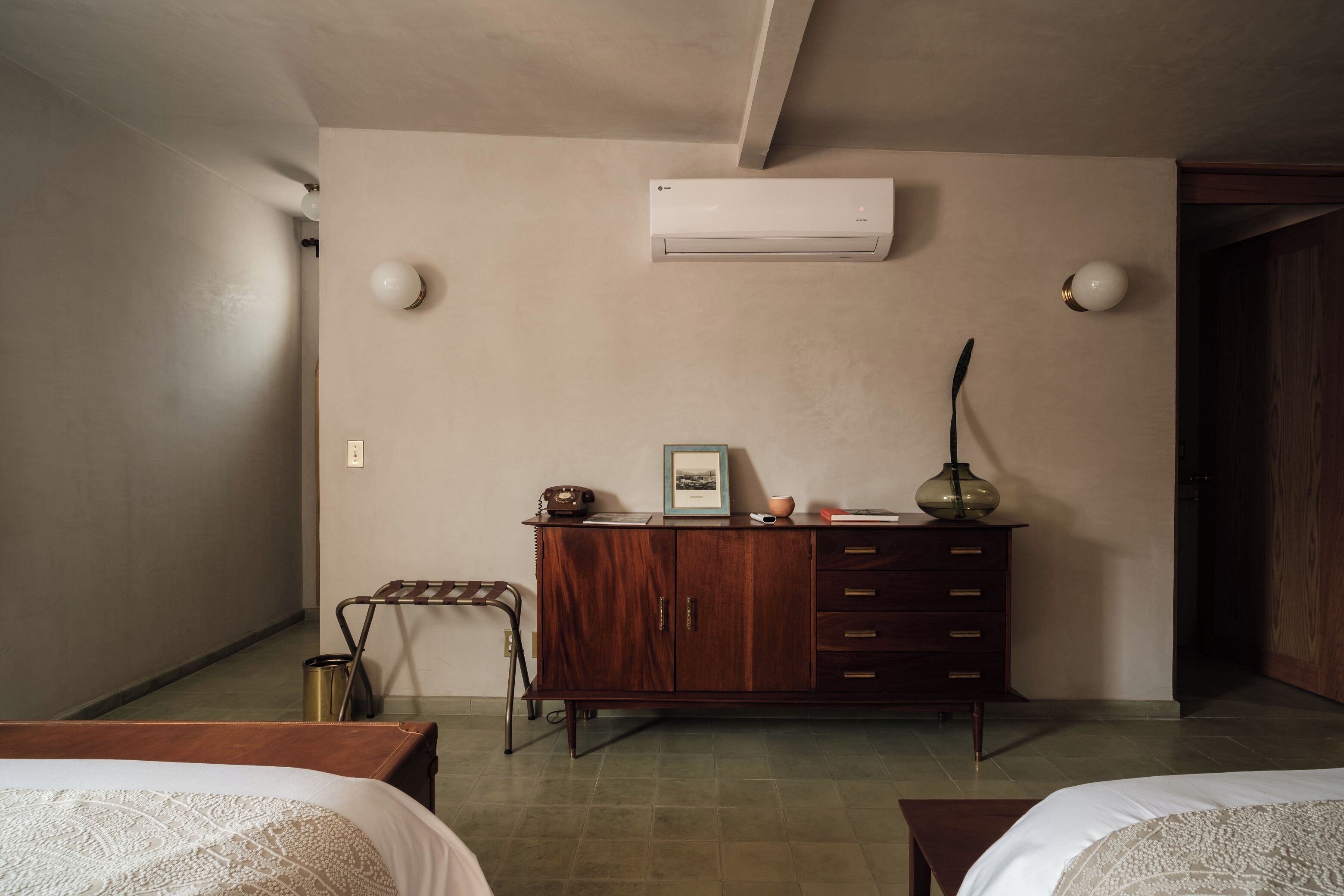 A hotel room with a bed, a wooden dresser, an air conditioner, and a small table.