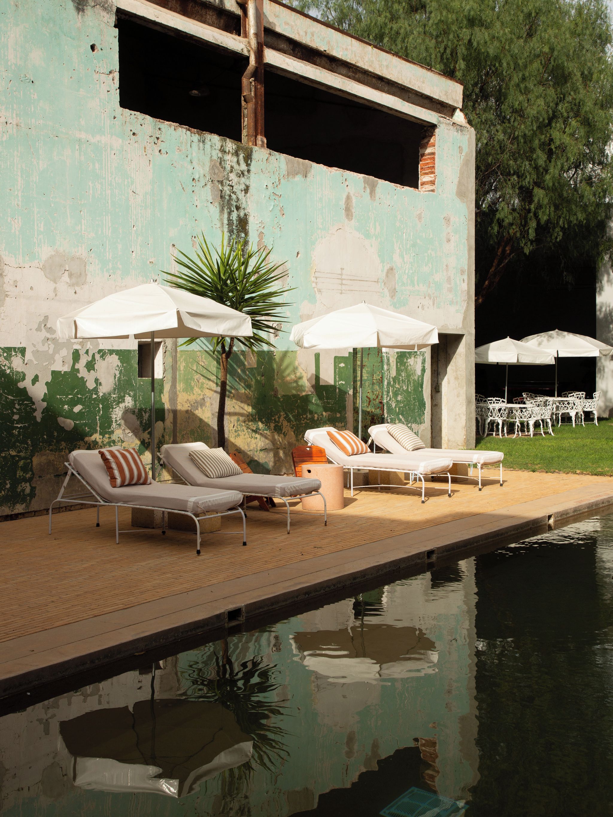 Un área junto a la piscina con sillones de descanso y sombrillas, una estructura de concreto con pintura descascarada y un reflejo en el agua.