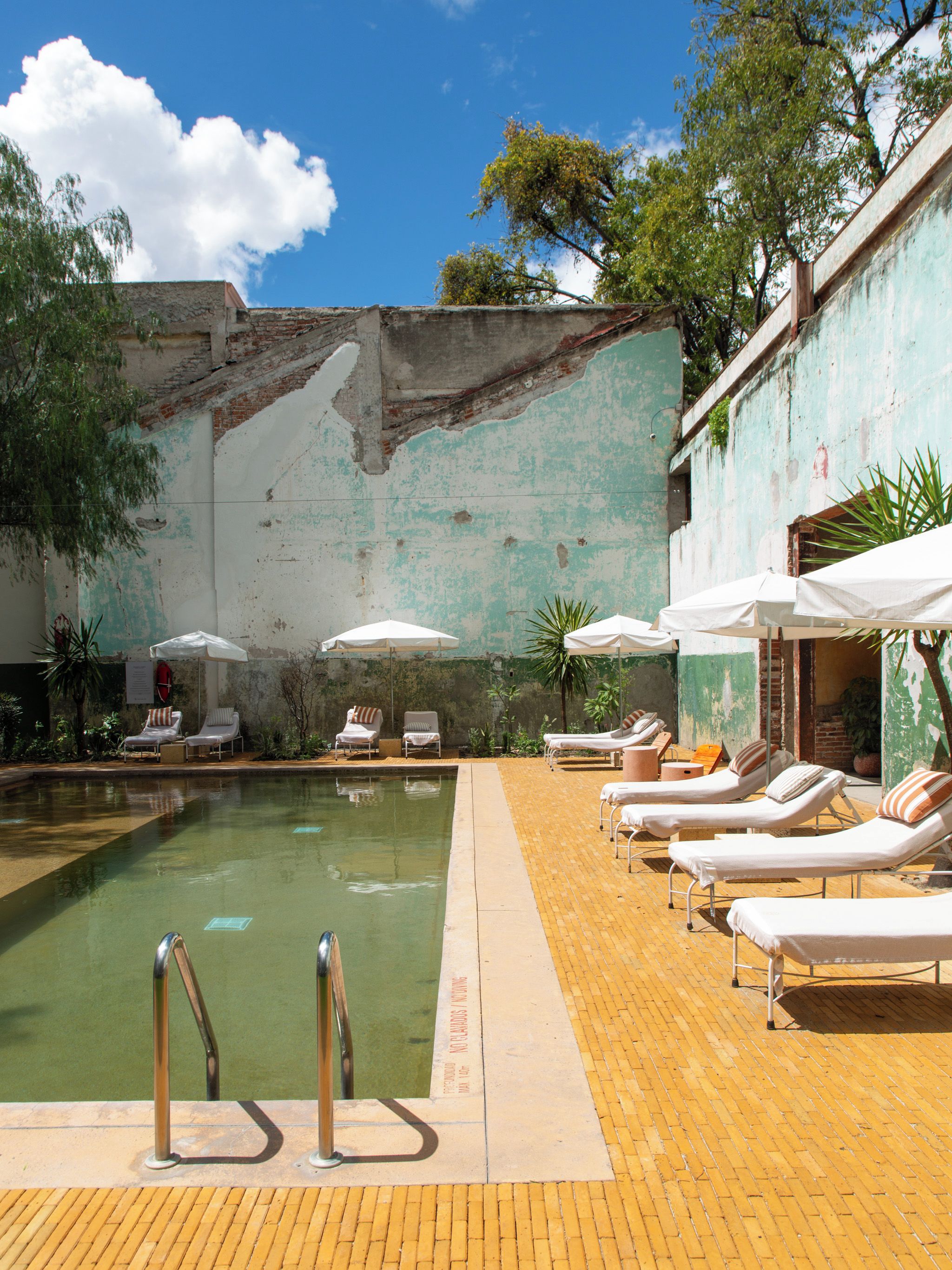A swimming pool with lounge chairs and umbrellas, surrounded by a brick wall with peeling paint.