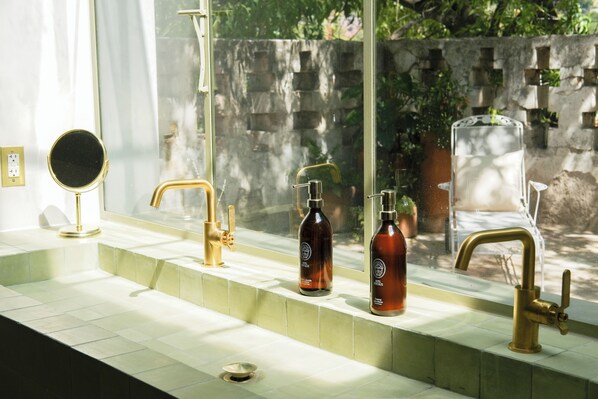 Un baño con lavamanos de azulejos blancos, grifo dorado, espejo redondo y dos botellas de jabón.