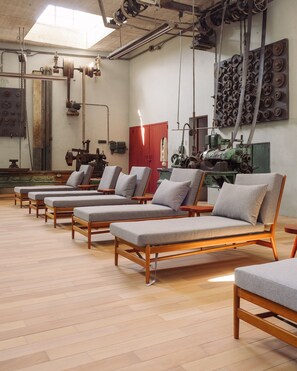A room with wooden flooring, multiple lounge chairs with grey cushions, and industrial-style machinery in the background. - Hotel Hércules (Querétaro)