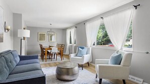 Ein modernes Wohnzimmer mit einem grauen Sofa, weißen Sesseln, einem Esstisch mit Stühlen und einem großen Fenster mit transparenten Vorhängen.