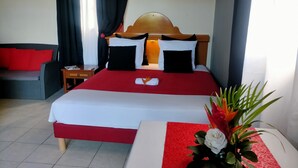A hotel room with a large bed, a wooden headboard, a red and white bedspread, a small table with a white tablecloth, a red and white runner, and a potted plant with a white flower. - Topaze Orientale Équipé Pour 2 Personnes Discret Sans Vis-à-vis! Vue sur Jardin! (Sainte-Rose)