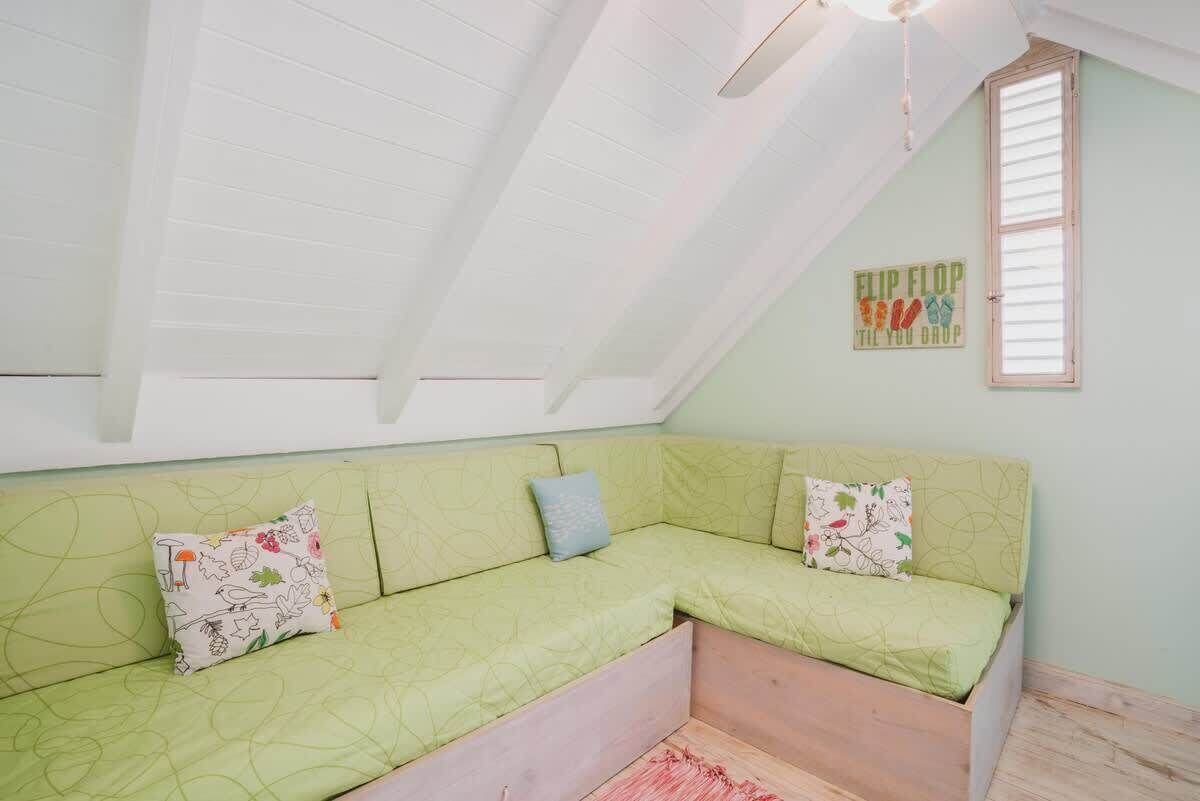 A room with a green sectional sofa, a window with blinds, and a ceiling fan.