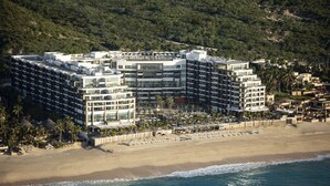 Et moderne hotel i flere etager beliggende ved siden af en strand med palmer og en klippebakke i baggrunden.
