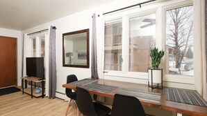 A modern room with a wooden dining table, black chairs, a TV on a stand, a large mirror, and a potted plant by the window. - Groundfloor Modern Industrial 2BD Apartment (Anchorage)