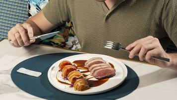 Una persona sosteniendo un cuchillo y un tenedor, a punto de comer un plato de carne en rodajas con salsa.