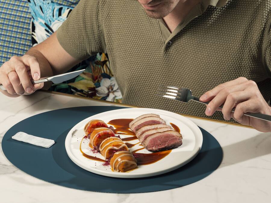 Una persona sosteniendo un cuchillo y un tenedor, a punto de comer un plato de carne en rodajas con salsa.