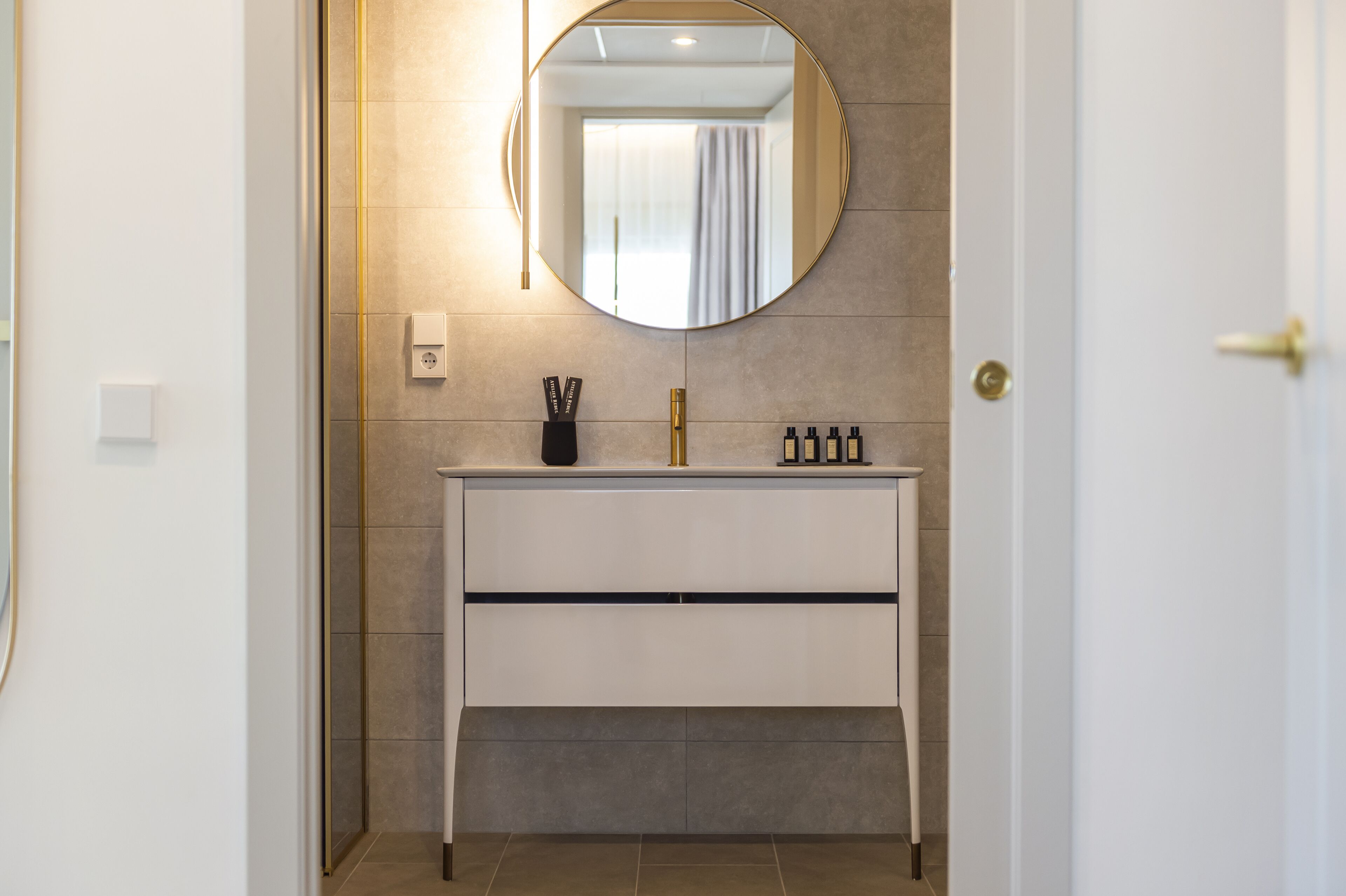 A modern bathroom with a white vanity, a round mirror, and a gold faucet.