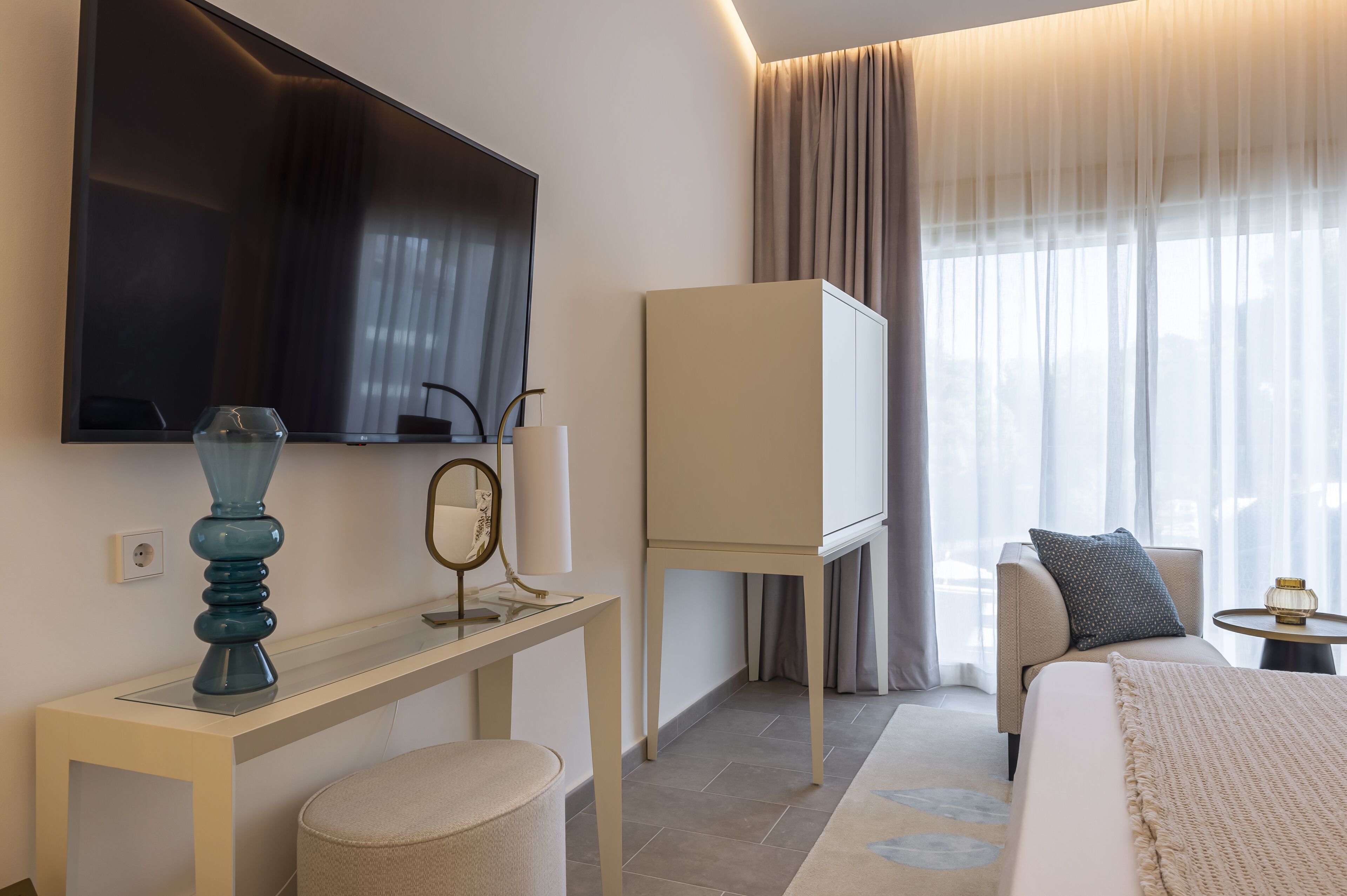 Una habitación de hotel moderna con un televisor de pantalla plana grande, una mesita de noche con lámpara, una pequeña mesa redonda con lámpara, un sofá con un cojín y un ventanal con cortinas translúcidas.