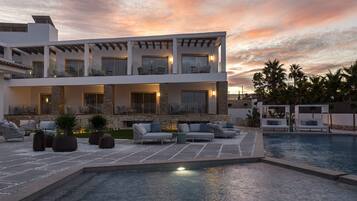 A modern two-story building with a pool, outdoor seating, and palm trees.