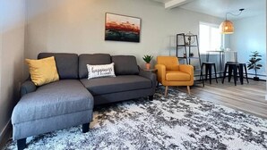 A living room with a grey sectional sofa, a yellow armchair, a black and white rug, a painting on the wall, and a dining area with a table and chairs. - Delightful Sleek Modern 2 Bed 1 Bath apt (Anchorage)
