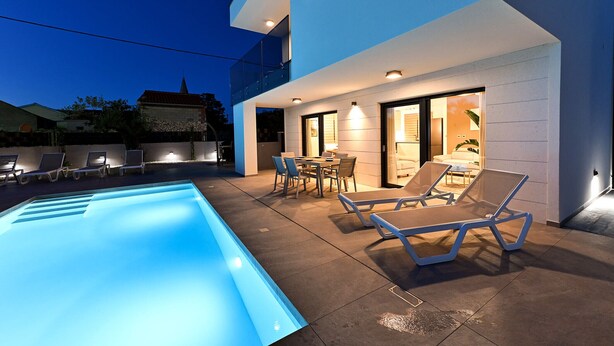 A modern outdoor pool area with lounge chairs and a dining table, surrounded by a two-story building with large windows and a visible balcony. - Villa Nostro Porto with heated pool (Kožino)