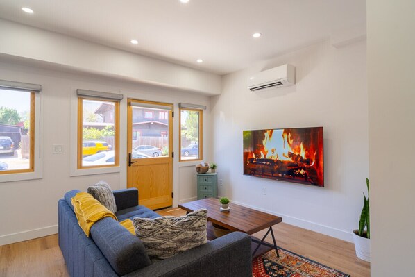 A modern living room with a sofa, a coffee table, and a painting of a fire. There are windows with blinds, a door, and an air conditioning unit on the wall.