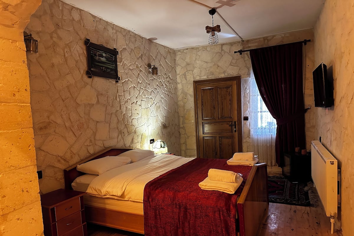 A bedroom with a stone wall, wooden bed, a red blanket, and a wooden dresser.