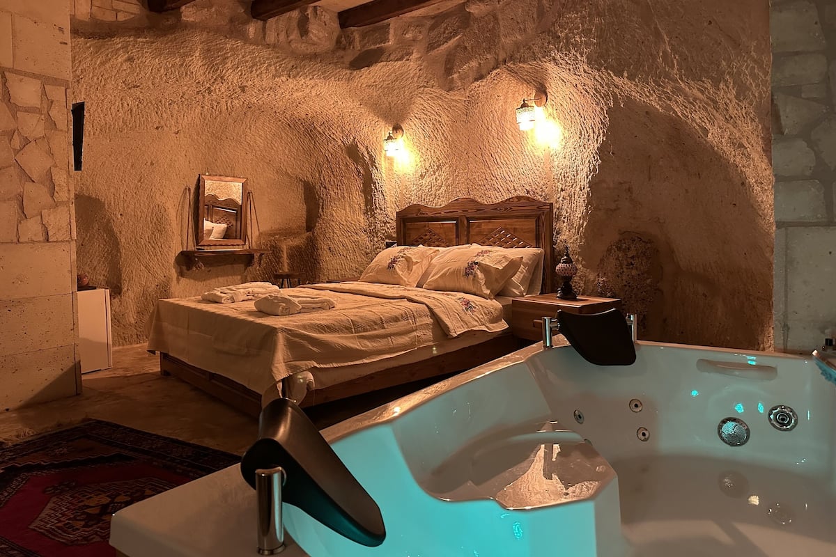 A rustic room with a stone wall, a wooden bed, a small table, and a bathtub.