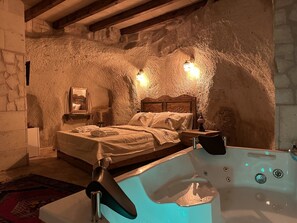 A rustic room with a stone wall, a wooden bed, a small table, and a bathtub. - Kale House Nar (Nevsehir)
