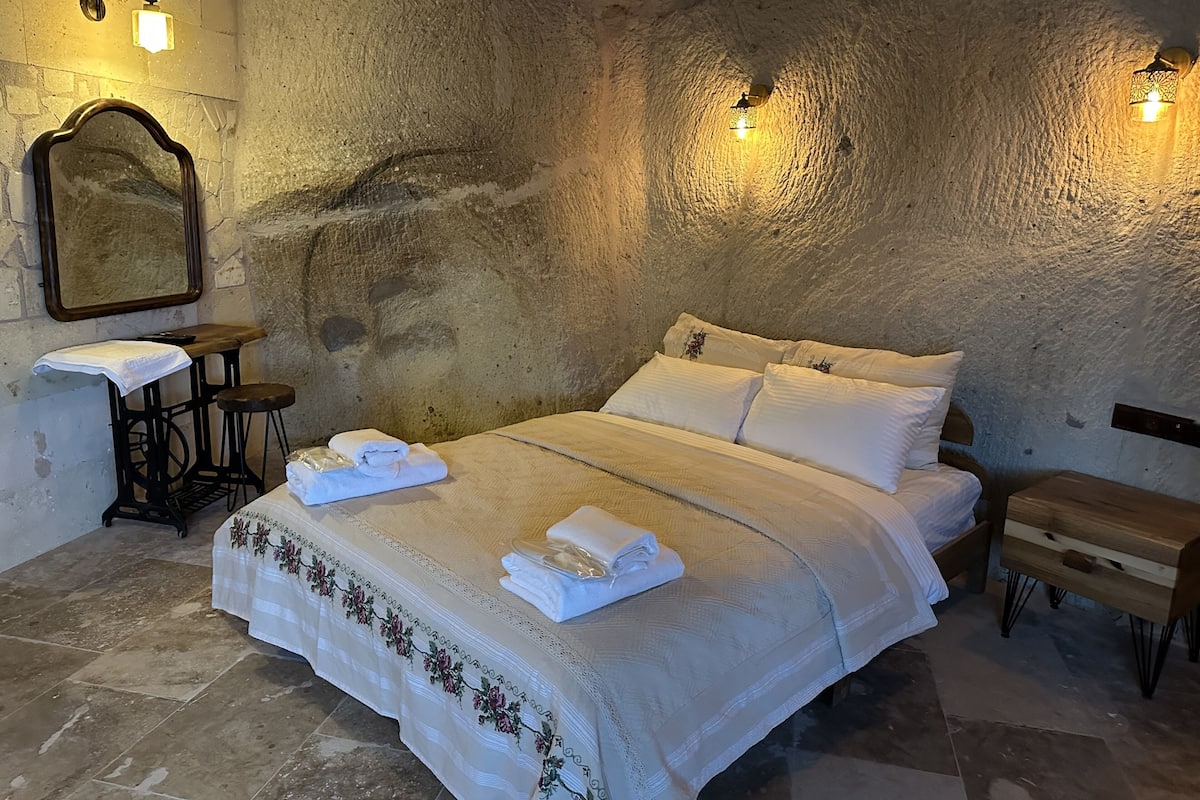 A bedroom with a stone wall, a bed with white linens, two nightstands, a wooden bench, and wall-mounted lights.
