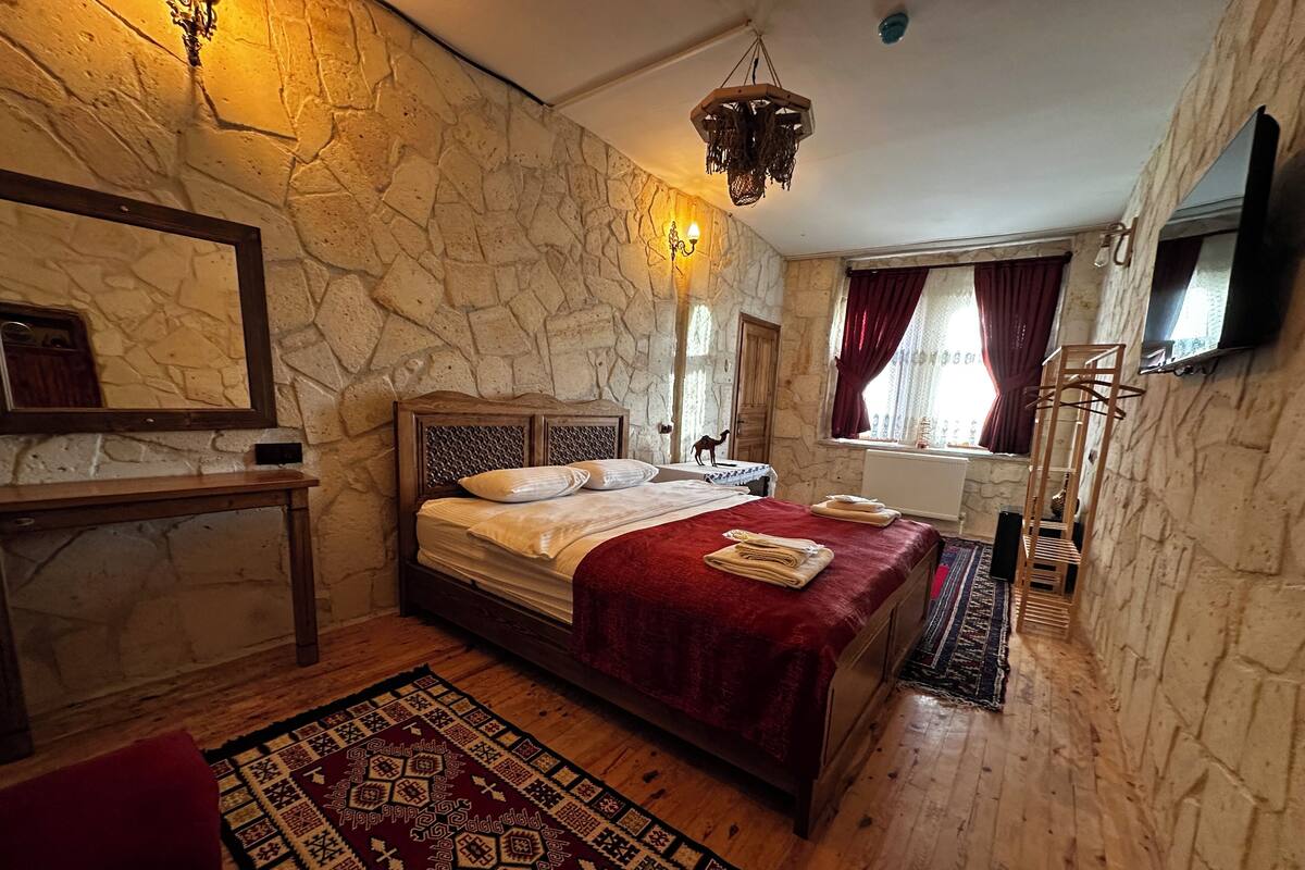 A bedroom with a stone wall, a wooden bed, a red blanket, a patterned rug, and a chandelier.