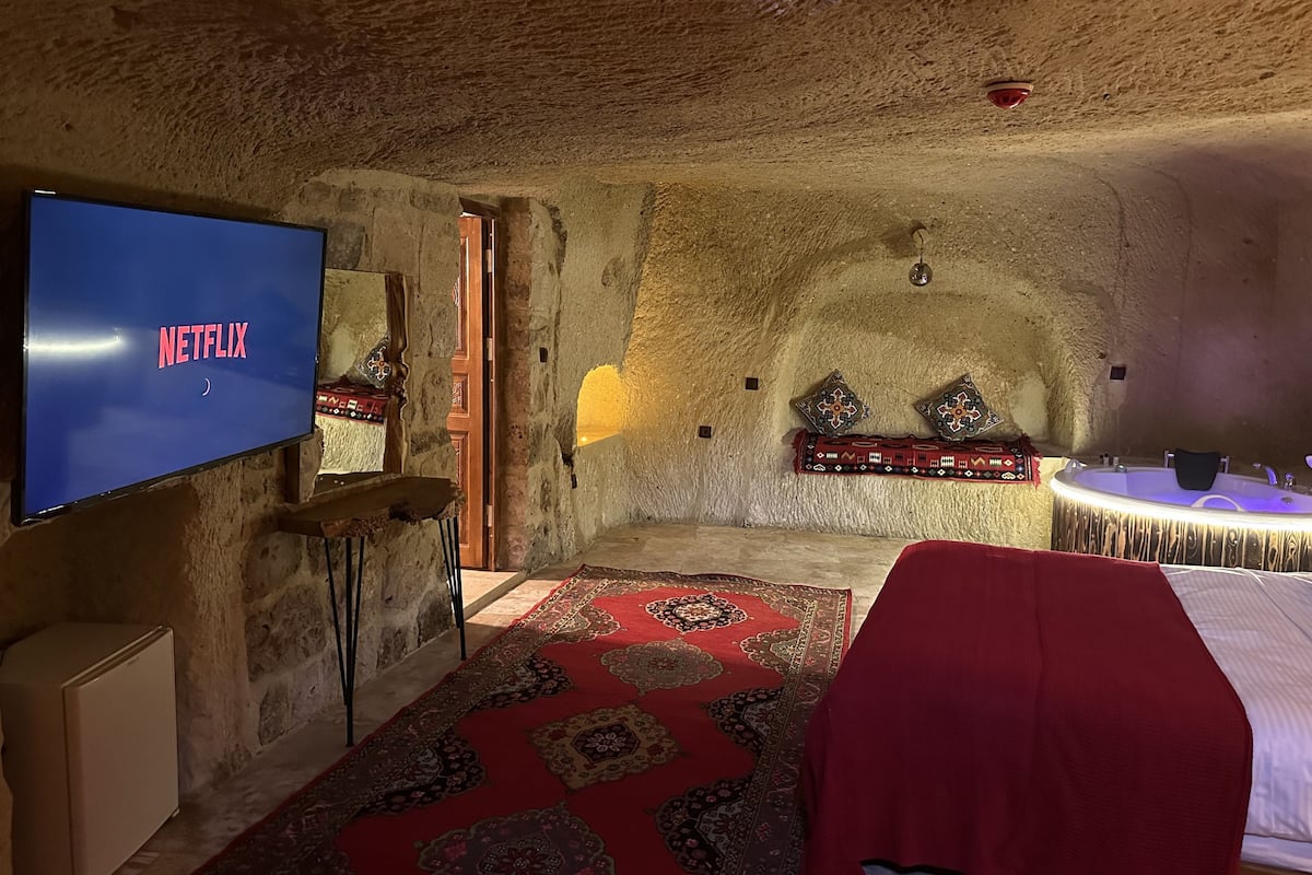 A cozy room with a stone ceiling, a flat-screen TV displaying 'NETFLIX', a large rug, a bathtub, and a small table with decorative pillows.