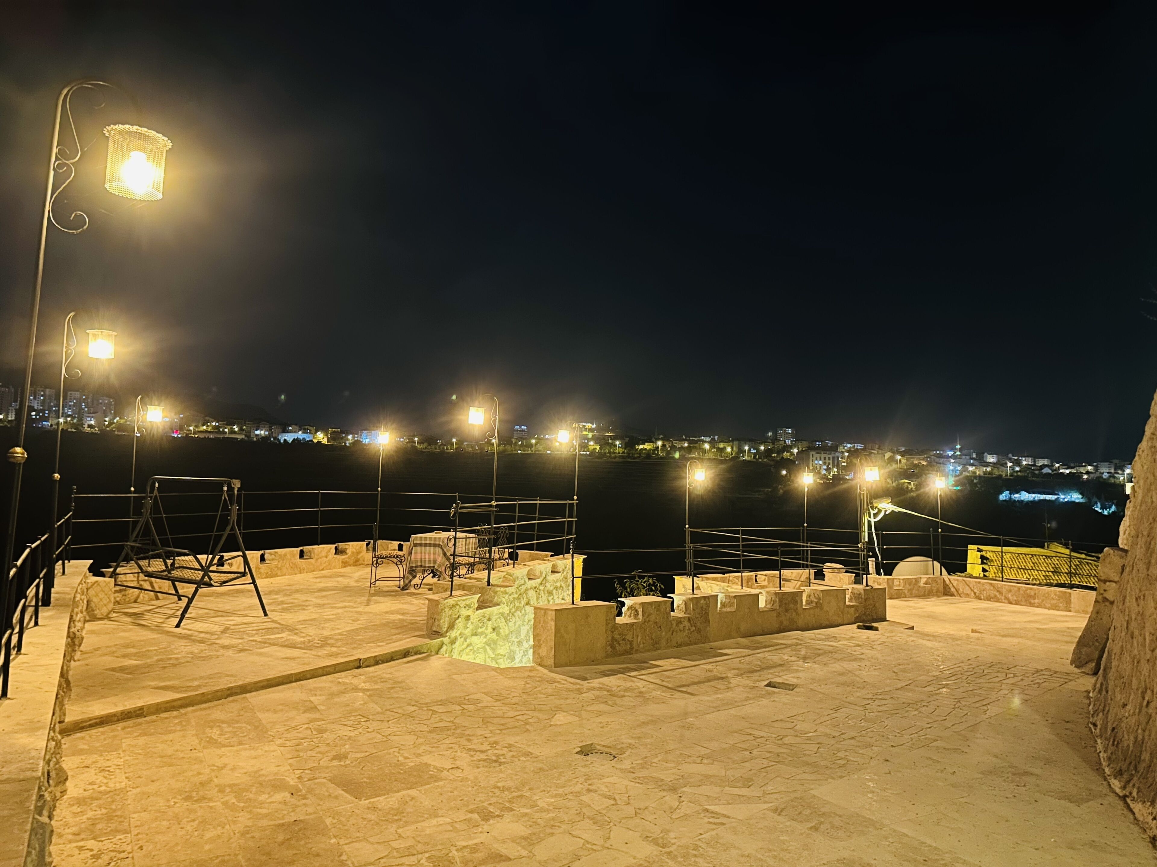 A rooftop terrace with a view of a cityscape at night, featuring street lamps and a railing.
