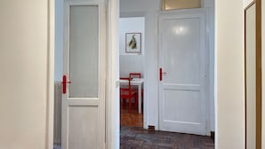 Ein Korridor mit weißen Türen, eine davon mit einem Milchglasfenster und roten Griffen, die andere schlicht und ebenfalls mit rotem Griff. Im Hintergrund sind ein roter Stuhl und ein Tisch zu sehen.