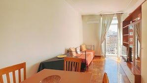 Ein Zimmer mit Esstisch, Stühlen, einem Bett, einem Fernseher und Blick auf eine Stadtlandschaft durch große Fenster.