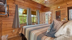 Une chambre à coucher dans une cabane en bois, avec un lit, des rideaux, un ventilateur de plafond et une fenêtre donnant sur un paysage verdoyant.