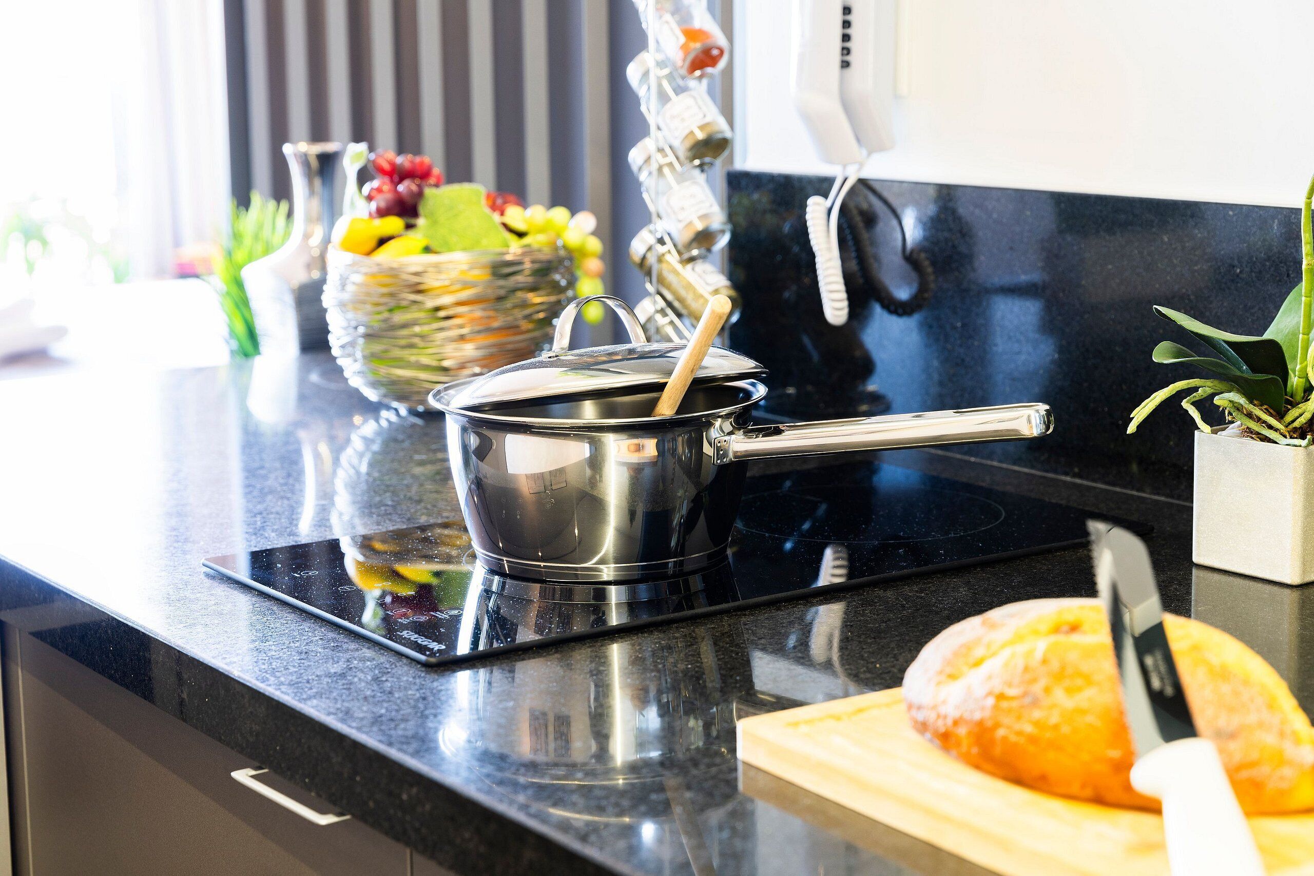Una encimera de cocina con una olla sobre una placa de inducción, una hogaza de pan en una tabla de cortar y una cesta de fruta.