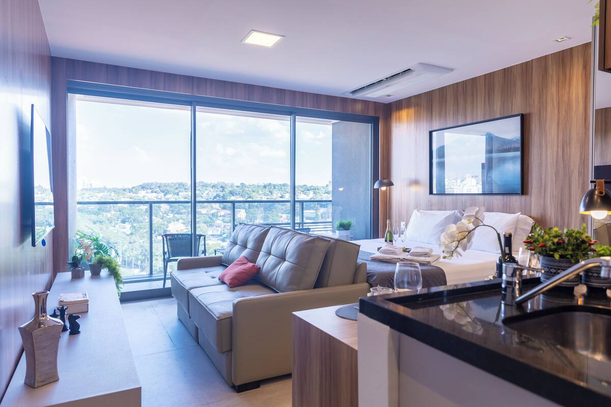 Ein modernes Hotelzimmer mit großem Fenster, einer Couch, einem Bett, einer Küchenzeile und Blick auf die Stadt.