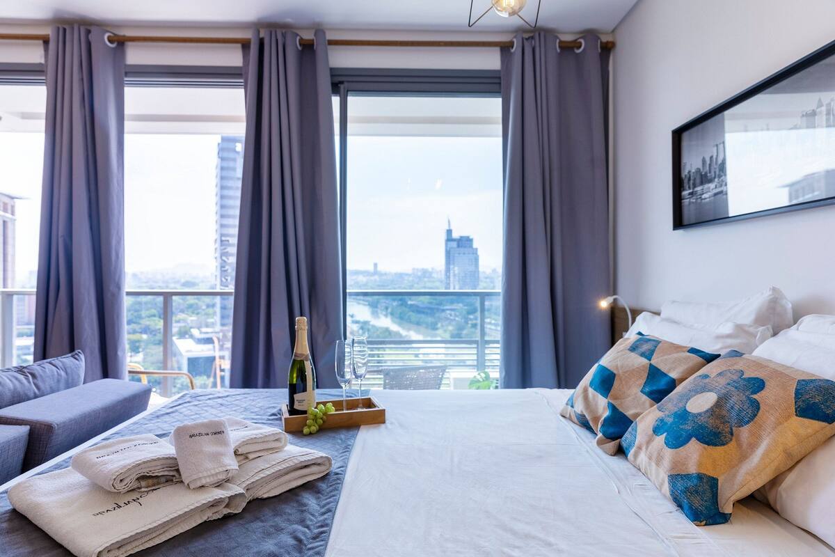 Ein Hotelzimmer mit Bett, Nachttisch, einem Fenster mit Vorhängen, Blick auf die Stadt und einem gerahmten Bild an der Wand.