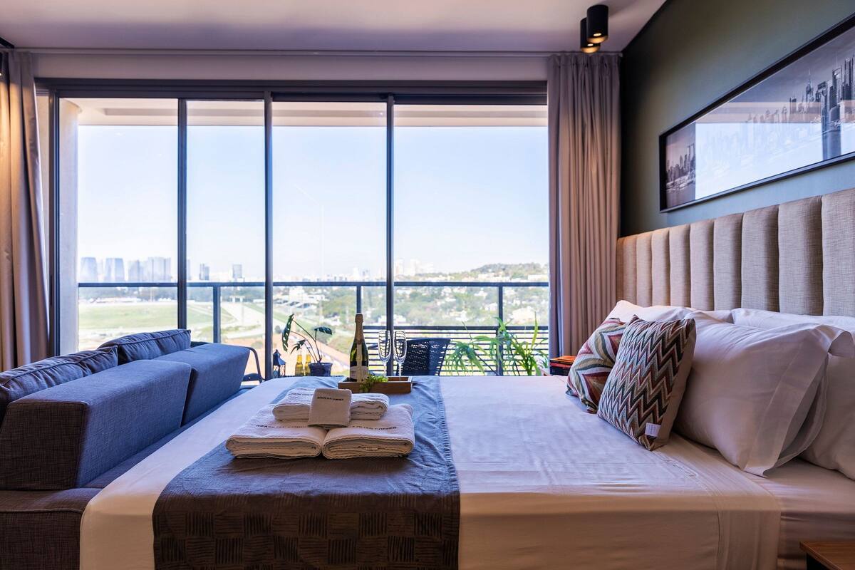 Ein modernes Hotelzimmer mit einem großen Bett, einer Sitzecke und Blick auf die Stadt durch raumhohe Fenster.