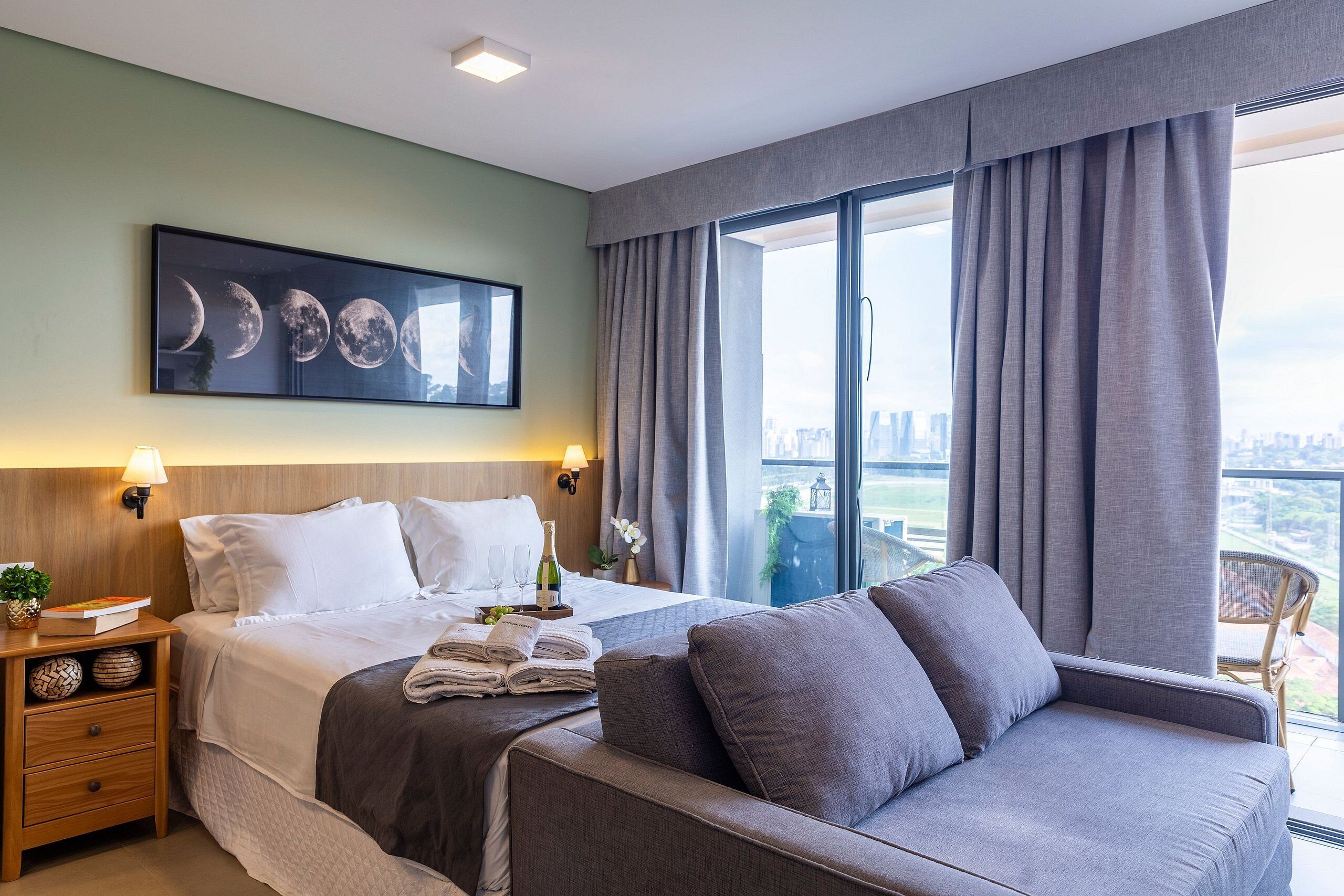 A hotel room with a bed, a sofa, a nightstand with a lamp, a framed picture of the moon phases, and a view of the city through large windows.