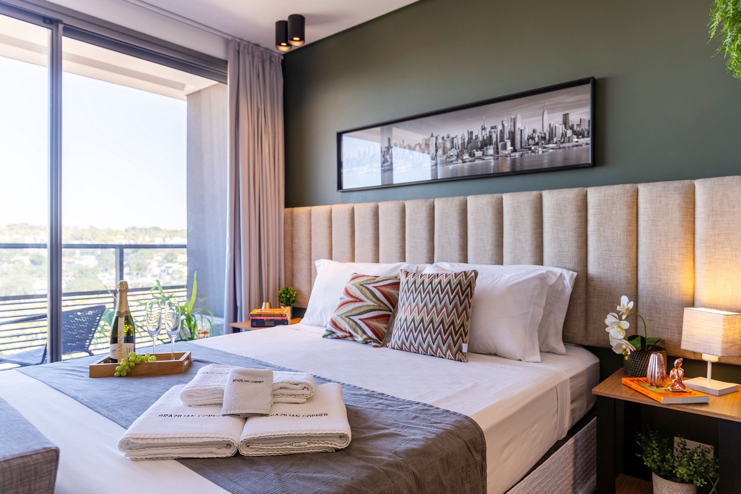 A modern hotel room with a large bed, a cityscape artwork on the wall, a balcony with a view, and a neatly made bed with towels and a bottle of champagne.