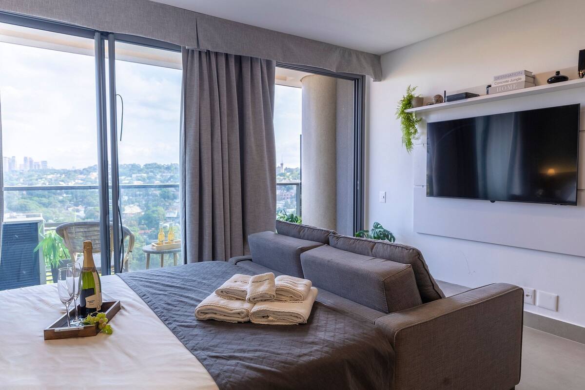 Ein modernes Hotelzimmer mit einem großen Bett, einer Couch, einem an der Wand befestigten Fernseher und Blick auf die Stadt durch das Fenster.