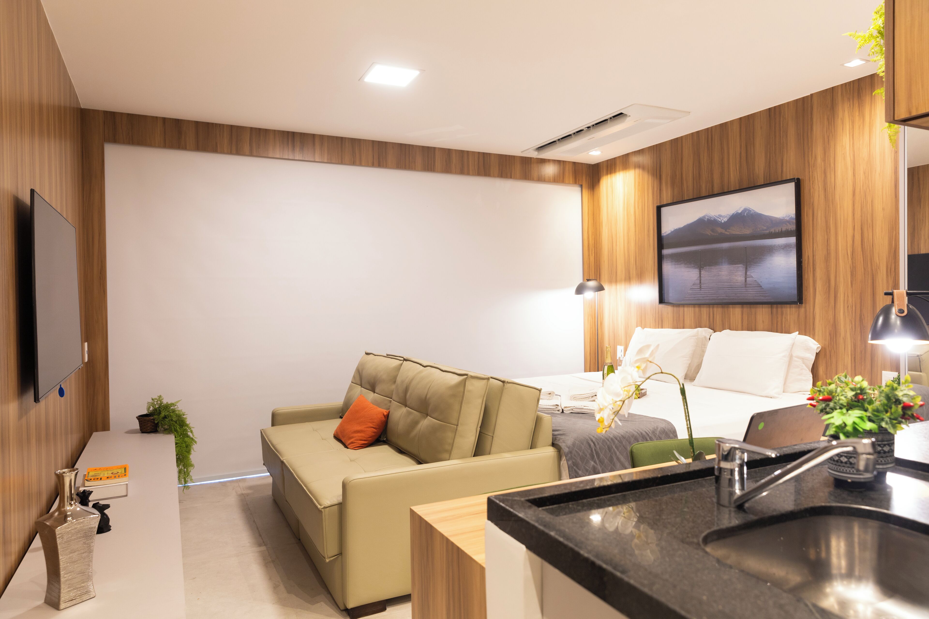 A modern hotel room with a sofa, a bed, a desk, and a kitchen area.