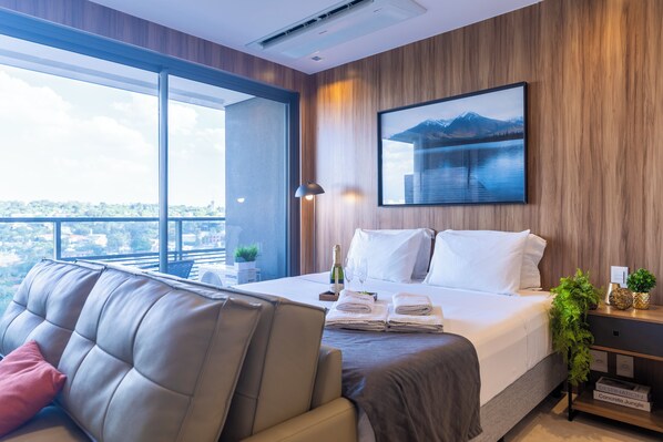 Moderni hotellihuone, jossa on suuri sänky, sohva, näköala kaupunkiin ja seinälle ripustettu taulu.