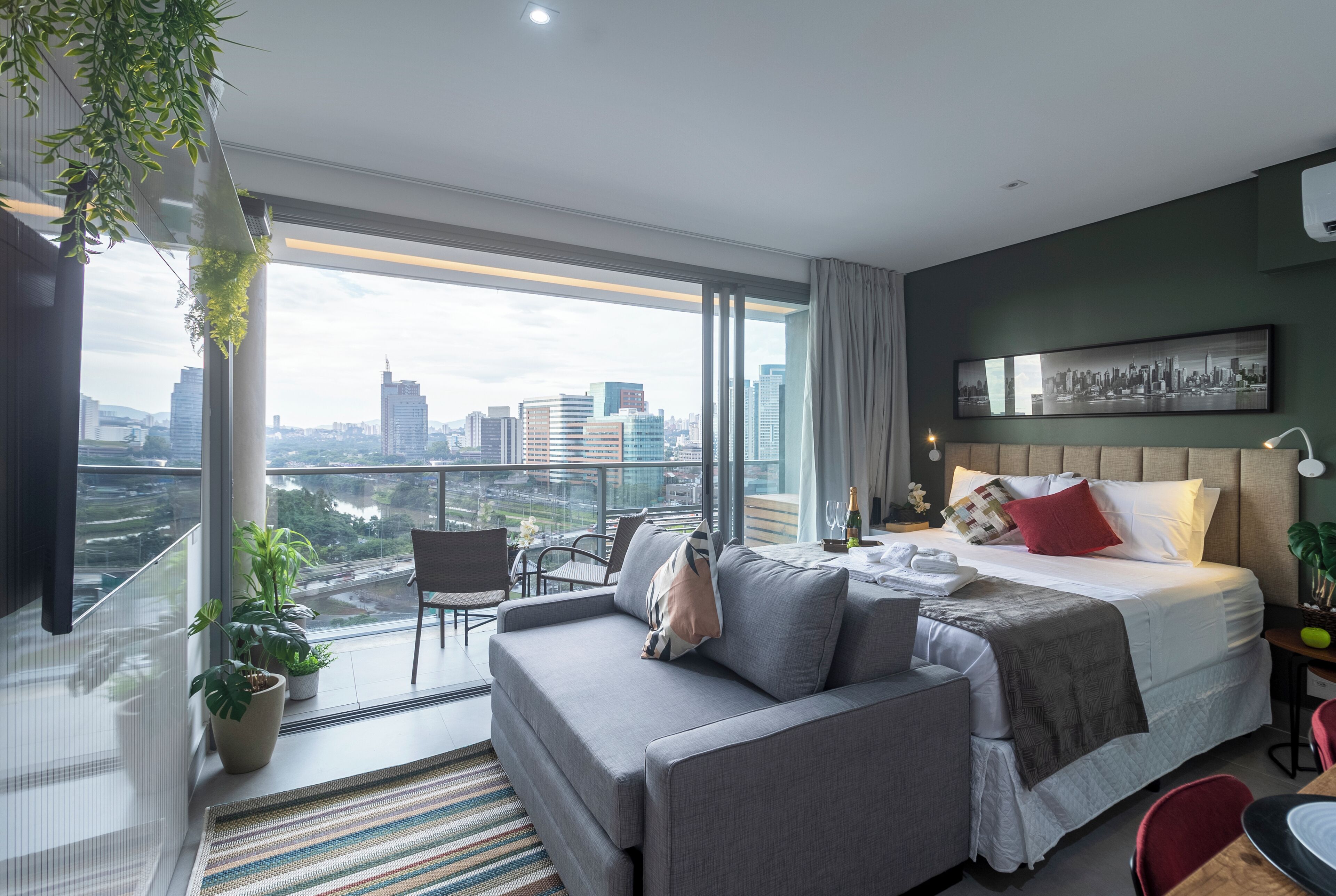 Una habitación de hotel moderna con un balcón grande, una cama con almohadas, un sofá y una zona de comedor.